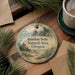 A round ornament with a scenic landscape of Bandon State, Oregon, featuring a lighthouse and a body of water. The ornament is hanging from a string and is placed on a wooden surface, surrounded by pine branches and a cup of coffee.