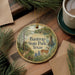 A decorative ornament with the text "Bastrop State Park, Texas" and an illustration of a deer in a forest setting, placed on a wooden surface alongside pine branches and a cup of coffee.