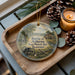 A decorative ornament with a scene depicting a wagon and a cabin, surrounded by pine cones and a lit candle, placed on a wooden tray.