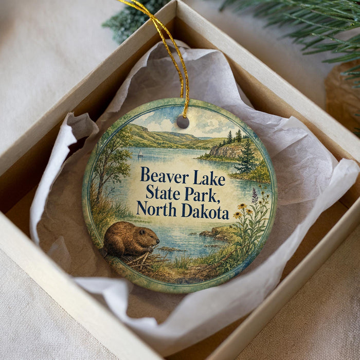 A round ornament with a beaver and a scenic landscape, placed in a box with a white tissue paper lining.