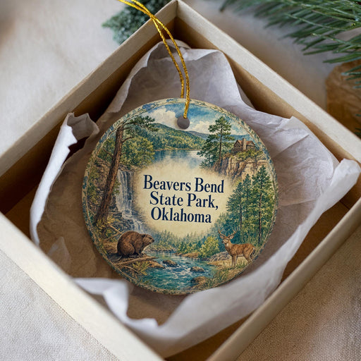 A circular ornament with a painting of a forest scene and a waterfall, placed in a box with a white tissue paper lining.