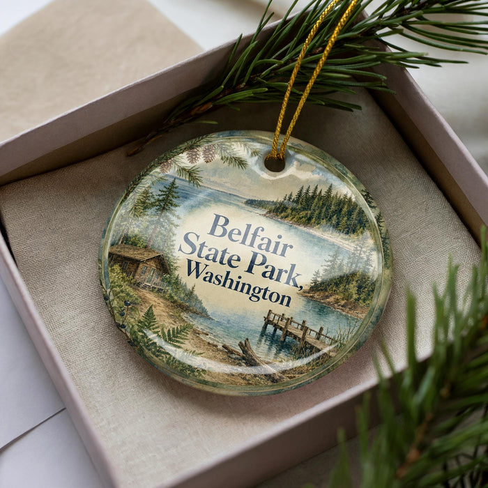 A decorative ornament in a box, featuring a scenic landscape with a lake, pine trees, and a cabin.