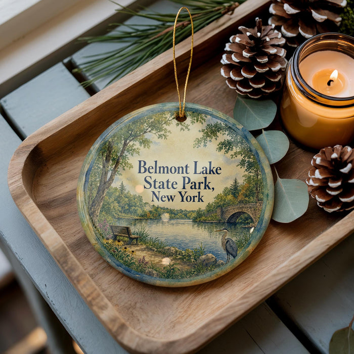 A round ornament with a painting of Belmont Lake State Park, New York, sits on a wooden tray next to a lit candle and pine cones.