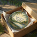 A decorative ornament in a box, featuring a scenic landscape with a lake, trees, and a bridge.