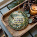 A round ornament with a scenic image of Belzville State Park, Pennsylvania, is displayed on a wooden tray alongside pine cones and a lit candle.