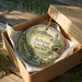 A decorative ornament in a box, featuring a scenic landscape with a bird and a building, with the text "Bethpage State Park, New York" printed on it.