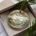 A decorative ornament in a box, featuring a scenic landscape of mountains, a river, and desert vegetation.