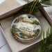A decorative ornament in a box, featuring a scenic landscape with a waterfall, pine trees, and a deer.