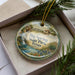 A decorative ornament in a box, featuring a scenic landscape with a waterfall, a house, and flowers.
