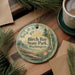 A round ornament with the text "Birch Bay State Park, Washington" is displayed on a wooden surface, surrounded by pine branches and a cup of coffee.