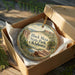 A decorative ornament in a box, featuring a scenic landscape with mountains and a river.