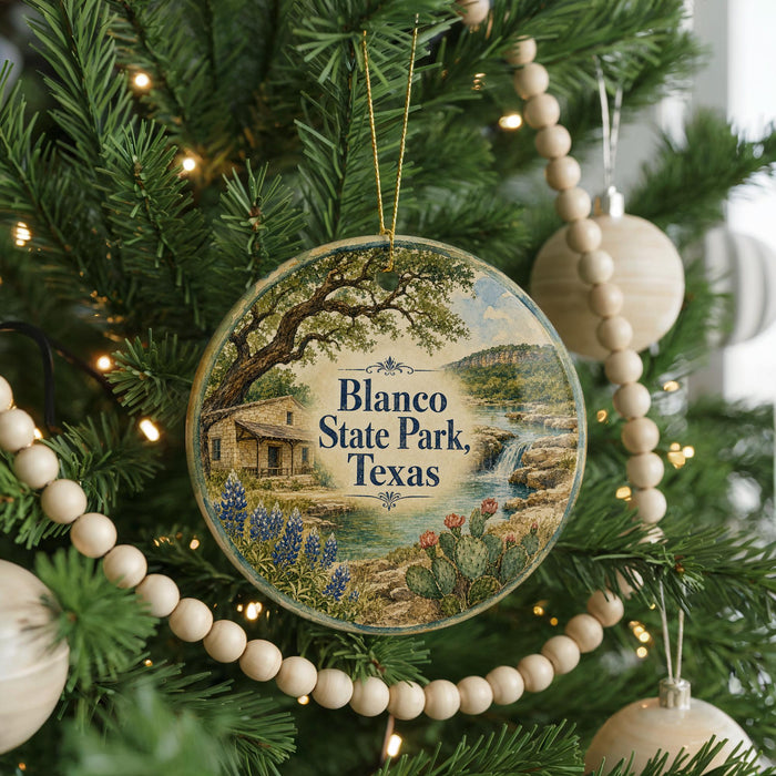 A circular ornament hanging on a Christmas tree, featuring a scenic landscape with a house, trees, and a waterfall.