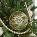 A circular ornament hanging on a Christmas tree, featuring a scenic landscape with a house, trees, and a waterfall.