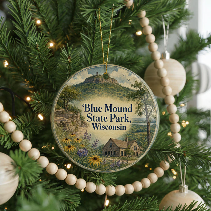 A round ornament hanging on a Christmas tree, featuring a scenic landscape with a mountain and a house, surrounded by greenery and flowers.