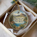 A decorative ornament in a white box, featuring a scenic landscape with mountains, trees, and a lake.