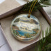 A decorative ornament in a box, featuring a scenic landscape with mountains, water, and greenery.