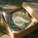 A decorative ornament in a box, featuring a scenic landscape with a river, bridge, and trees.