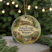 A round ornament with a painting of a lodge and a river, surrounded by greenery, hangs in front of a blurred background with twinkling lights.