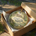 A round ornament in a wooden box, featuring an illustration of a historic lodge surrounded by trees and a stream.
