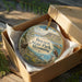 A circular ornament in a box, featuring a scenic landscape with mountains, water, and wildlife, including a bird and a deer.
