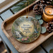 A decorative ornament with a scenic image of Breaks Interstate Park, Virginia, featuring a waterfall and a gazebo, placed on a wooden tray alongside pine cones and a lit candle.