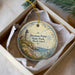 A round ornament with a lighthouse and coastal scene, placed in a box with a white tissue paper lining.