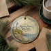 A round ornament with a lighthouse and coastal scene, placed on a wooden surface next to a cup of coffee and some pine branches.