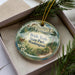 A decorative ornament in a box, featuring a scenic image of the Bridalle Trails State Park in Washington.