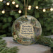 A circular ornament with a scenic landscape and wildlife, featuring a cabin, trees, and a bird, hangs in front of a Christmas tree with lights.