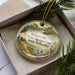A decorative ornament with a landscape scene and text, placed in a box with pine branches.