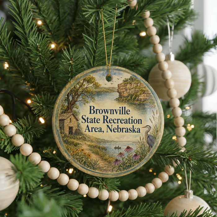 A round ornament hanging on a Christmas tree, featuring a scenic landscape with a house, trees, and a bird.
