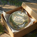 A circular ornament in a box, featuring a scenic landscape with a waterfall and forest, with the text "Bucktail State Park, Pennsylvania" printed on it.