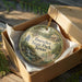 A decorative ornament in a box, featuring a scenic landscape with a house and trees, with the text "Buescher State Park, Texas" printed on it.