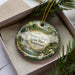A decorative ornament with a scenic image of a waterfall and forest, placed in a box with pine branches.