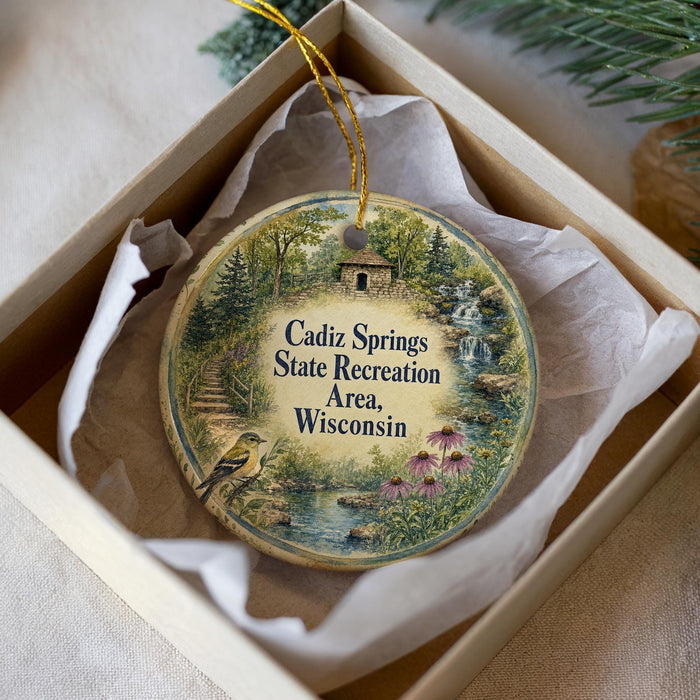A decorative ornament in a white box, featuring a scenic landscape with a bird, trees, and a waterfall.