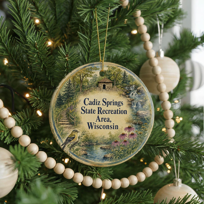 A round ornament hanging on a Christmas tree, featuring a scenic landscape with a bird and a house.