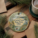 a decorative ornament with a scenic landscape of Caesar Creek State Park, Ohio, surrounded by pine branches and a cup of coffee on a wooden surface.