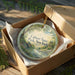 A decorative ornament in a box, featuring a scenic landscape with a river and trees, with the text "Calvert Park, New York" printed on it.