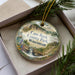 A decorative ornament in a box, featuring a scenic landscape with a waterfall, pine trees, and a bridge.