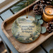 A decorative ornament with a serene landscape of Camano Island, Washington, featuring a heron, pine trees, and a body of water. The ornament is placed on a wooden tray, with pine cones and a lit candle nearby.