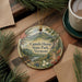 A decorative ornament with a scenic mountain landscape and text, placed on a wooden surface next to a cup of coffee and pine branches.