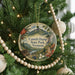 A round ornament on a Christmas tree, featuring a scenic landscape with mountains, pine trees, and a river.
