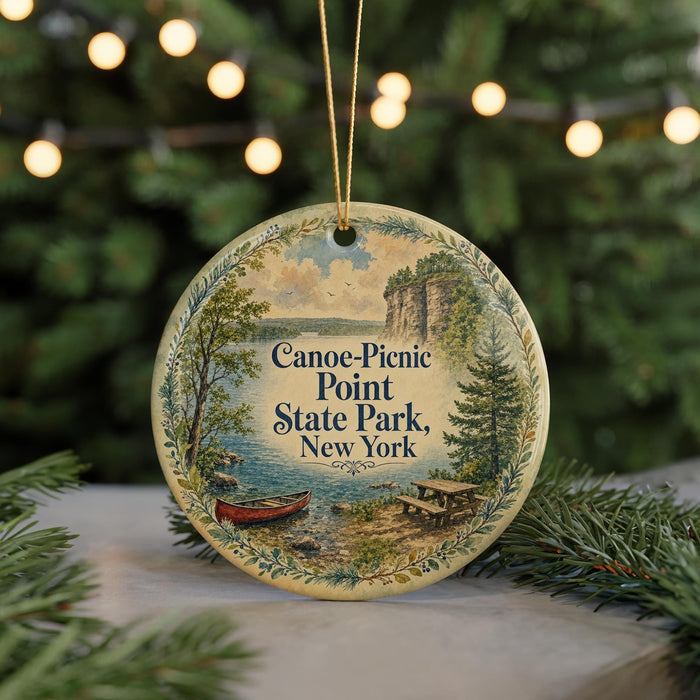 A round ornament with a painting of a canoe and picnic area, hanging in front of a Christmas tree with lights.