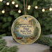 A round ornament with a painting of a canoe and picnic area, hanging in front of a Christmas tree with lights.