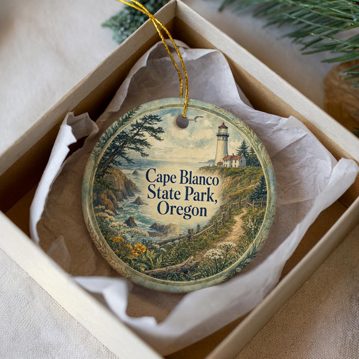 A decorative ornament in a box, featuring a lighthouse and a scenic landscape, with the text "Cape Blanco State Park, Oregon" printed on it.