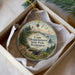 A round ornament with a lighthouse and a coastal landscape, placed in a box with a white tissue paper lining.