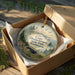 A round ornament in a box, featuring a scenic landscape with a lighthouse and mountains, with the text "Cape Disappointment State Park Washington" printed on it.