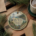 A round ornament featuring a lighthouse and a scenic view of Cape Meares State Scenic Viewpoint, Oregon, is displayed on a wooden surface alongside a cup of coffee and some pine branches.