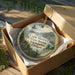 A decorative ornament in a box, featuring a lighthouse and a scenic view of Cape Meares State Scenic Viewpoint, Oregon.