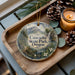 A decorative ornament featuring a scenic landscape of Cascadia State Park, Oregon, with pine cones and a lit candle on a wooden tray.